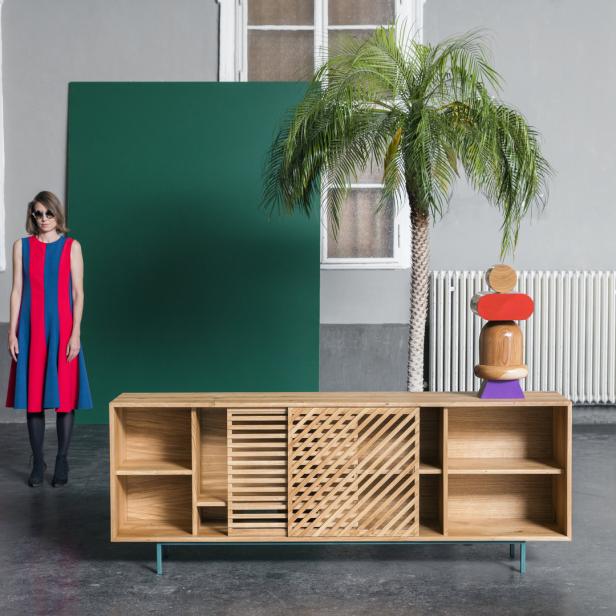 Eine Frau in einem rot-blau gestreiften Kleid steht neben einem Sideboard mit einer Palme darauf.