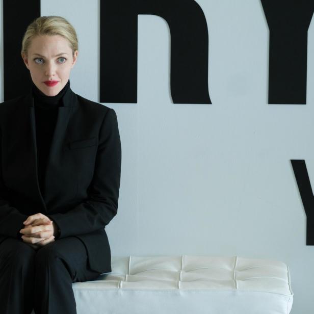 Amanda Seyfried sitzt in einem schwarzen Hosenanzug vor einer Wand mit schwarzen Buchstaben.