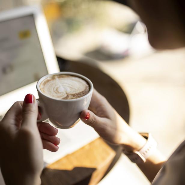 Eine Frau hält eine Tasse Cappuccino vor einem Laptop.