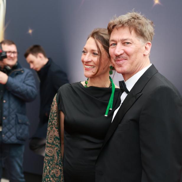 Ein Mann im Smoking und eine Frau in einem schwarzen Kleid posieren auf einem roten Teppich.