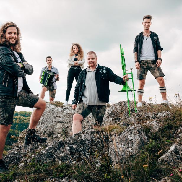 Eine Band in Lederhosen posiert mit Akkordeon und Posaune auf einem Felsen.