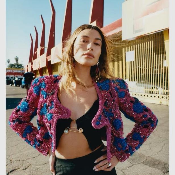 Hailey Bieber posiert in einer bunten Jacke und einem schwarzen Top vor einem Gebäude.