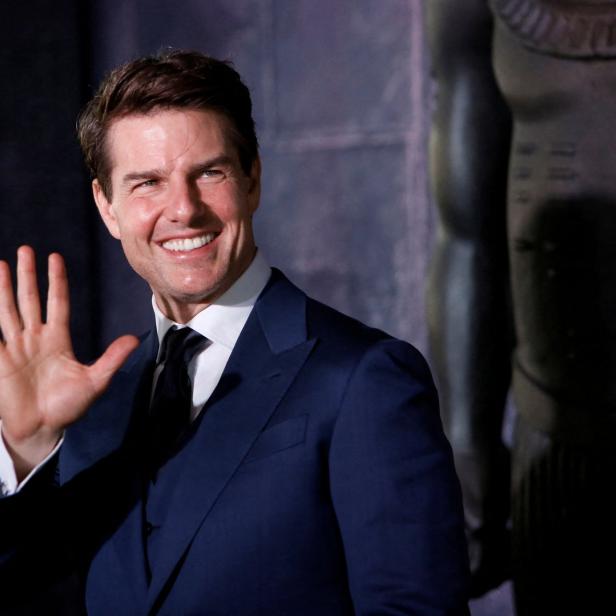 Tom Cruise lächelt und winkt in einem blauen Anzug.