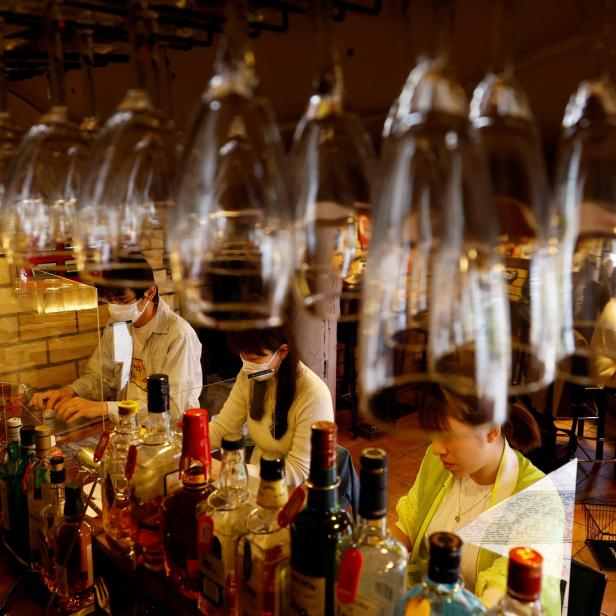 Drei Personen mit Masken arbeiten hinter einer Bar mit vielen Flaschen und Gläsern.