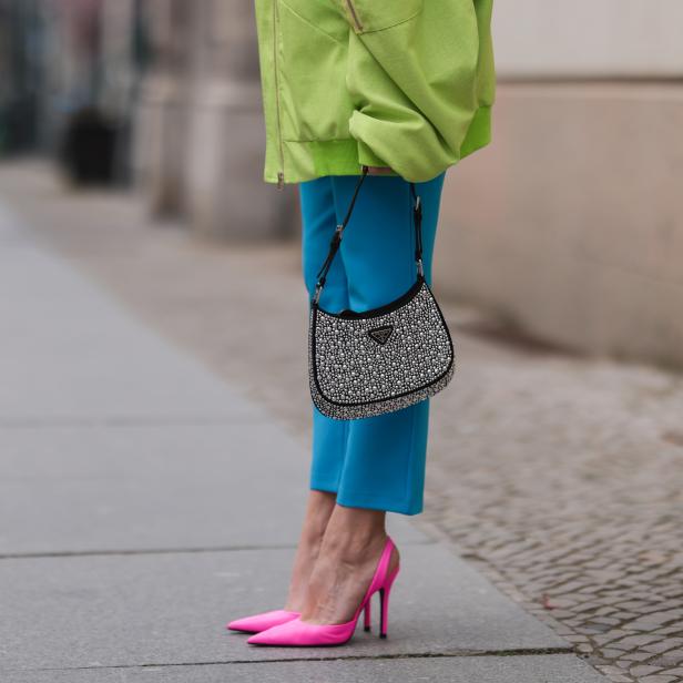 Nahaufnahme einer Person mit hellgrüner Jacke, blauen Hosen, einer Prada-Tasche und pinkfarbenen Schuhen.