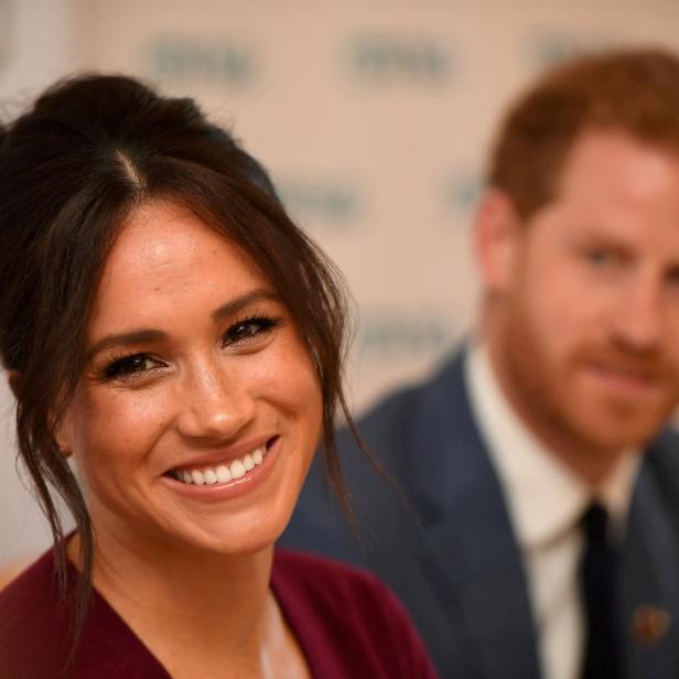 Meghan Markle lächelt, während Prinz Harry im Hintergrund zu sehen ist.