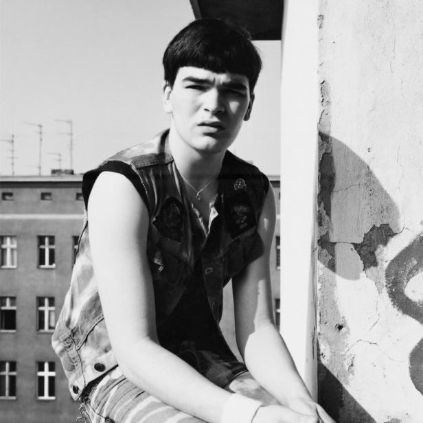 Ein junger Mann mit Bowlcutschnitt und Jeansjacke sitzt vor einem Gebäude.