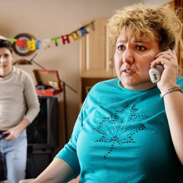 Eine besorgte Frau telefoniert, während zwei Jungen im Hintergrund stehen; eine Geburtstagsgirlande ist zu sehen.