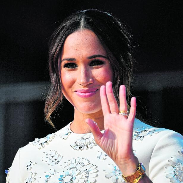 Meghan Markle lächelt und winkt in einem weißen Kleid mit floralen Verzierungen.