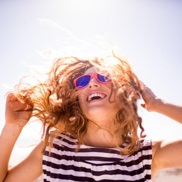Eine lachende Frau mit lockigen Haaren und pinkfarbener Sonnenbrille am Strand.