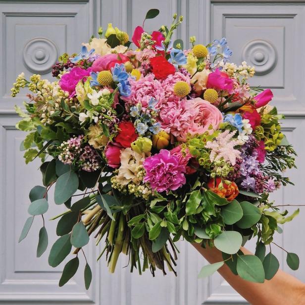 Ein bunter Blumenstrauß wird vor einer grauen Tür gehalten.