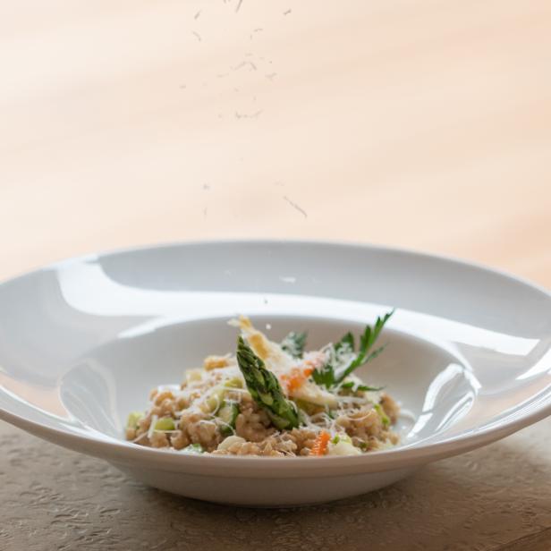 Eine Person reibt Käse über ein Gericht mit Spargel und Kräutern.