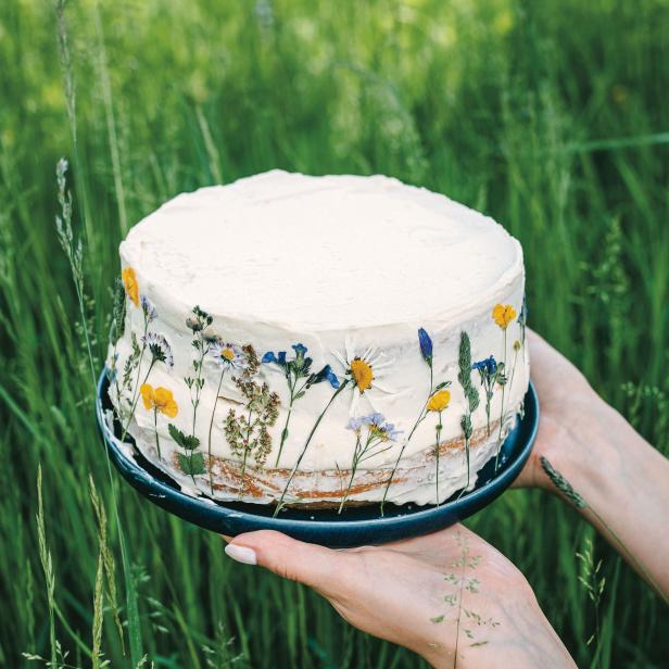 Ein mit essbaren Blumen dekorierter Kuchen wird von zwei Händen gehalten.