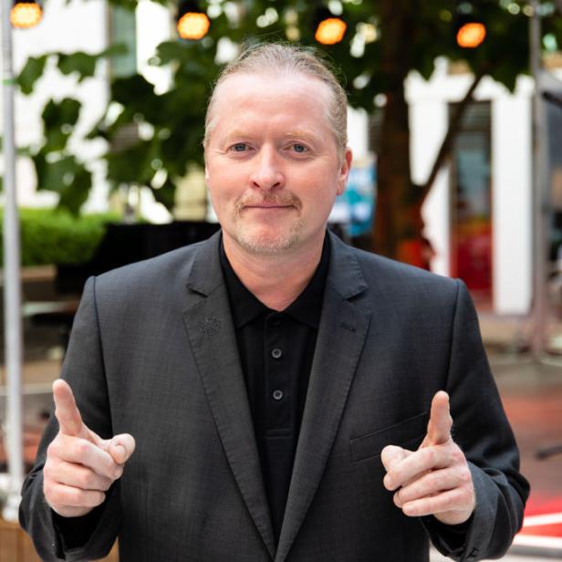 Ein Mann im Anzug zeigt mit beiden Händen auf die Kamera.
