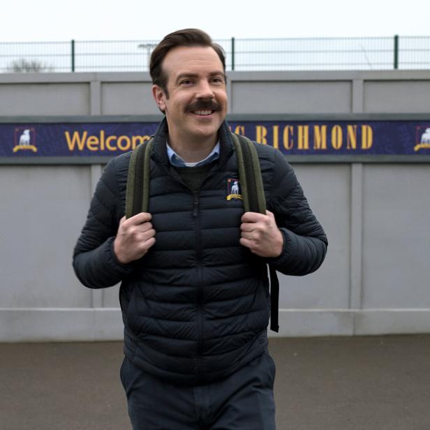 Ted Lasso mit Rucksack vor einem „Welcome to Richmond“-Schild.