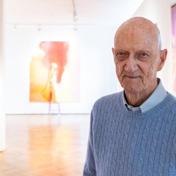 Ein älterer Mann steht in einer Kunstgalerie vor abstrakten Gemälden und einer lila Skulptur.