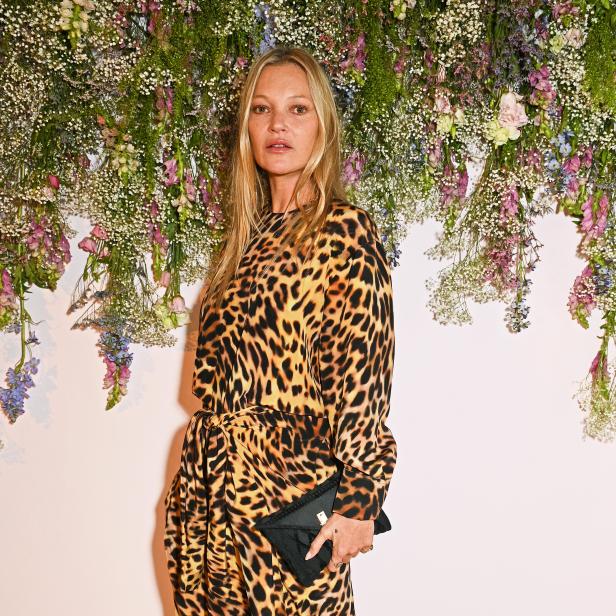 Kate Moss in einem Leopardenmuster-Overall mit einer schwarzen Clutch vor einem Blumenhintergrund.