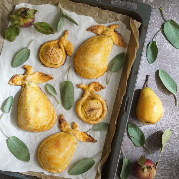Auf einem Backblech liegen gebackene Birnen- und Apfeltaschen mit Salbeiblättern.