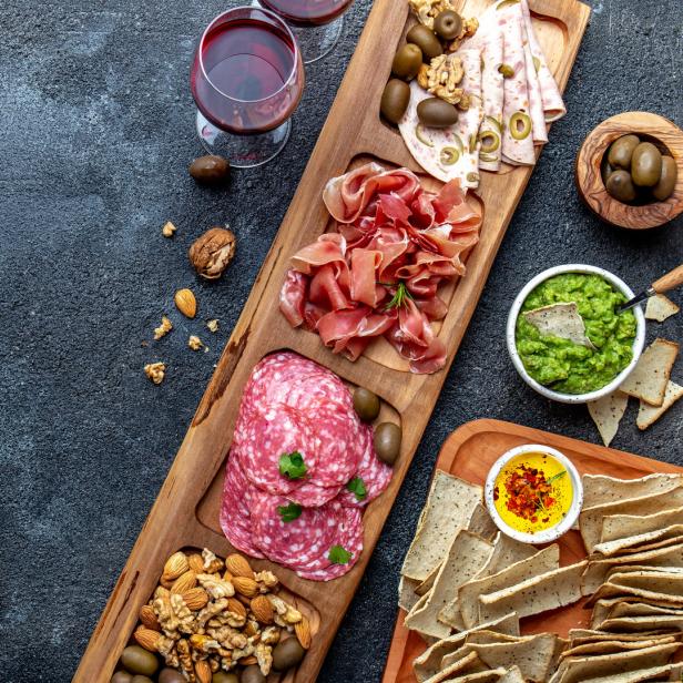 Eine Holzplatte mit Wurst, Käse, Nüssen, Oliven, Dips und zwei Gläsern Rotwein.