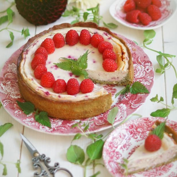 Ein Himbeerkuchen auf einem Teller mit floralem Muster, umgeben von Blumen und Blättern.