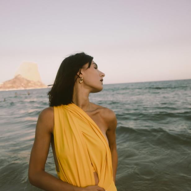 Eine Frau in einem gelben Kleid steht am Strand, im Hintergrund ein Felsen im Meer.
