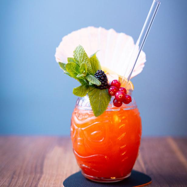 Ein Tiki-Cocktail mit Beeren, Minze und einer Muschel als Dekoration auf einem Holztisch.