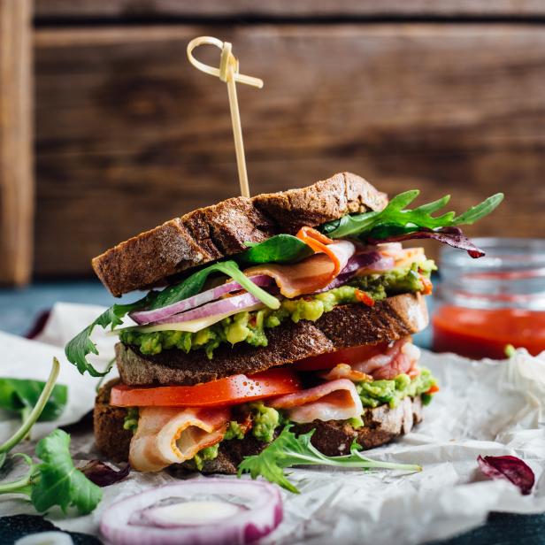 Ein aufgeschnittenes Sandwich mit Avocado, Speck, Tomaten und Salat auf dunklem Brot.
