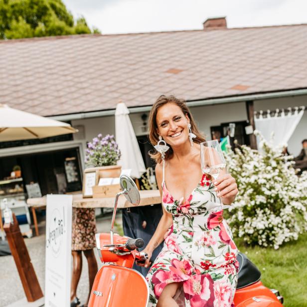 Eine Frau im Sommerkleid sitzt auf einer orangefarbenen Vespa und hält ein Weinglas in der Hand.