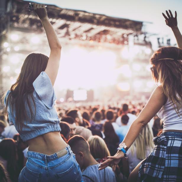 Zwei junge Frauen sitzen auf den Schultern von Freunden bei einem Konzert und jubeln.