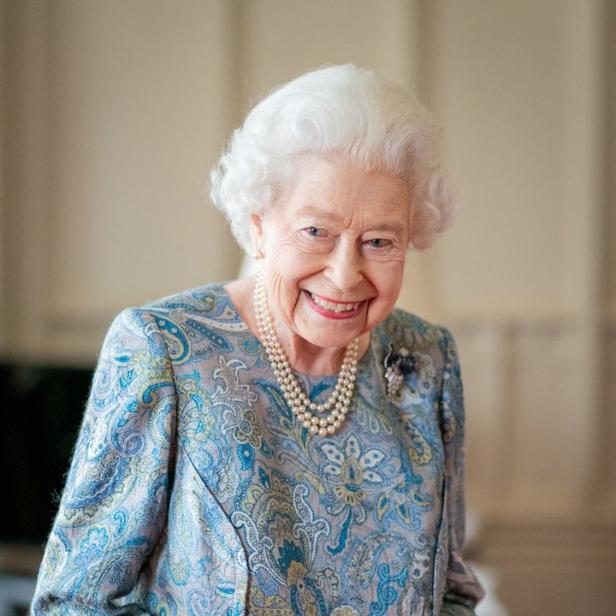 Königin Elizabeth II. lächelt in einem blau gemusterten Kleid und einer dreireihigen Perlenkette.