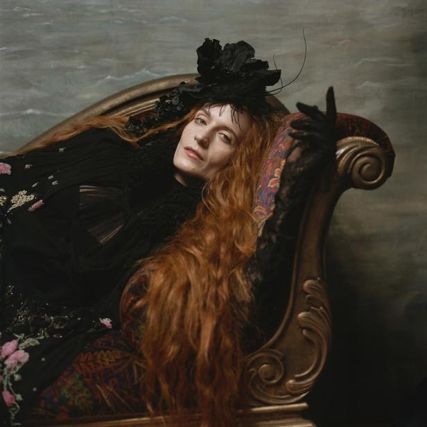 Florence Welch liegt in einem schwarzen Kleid und mit Hut auf einer verzierten Couch.