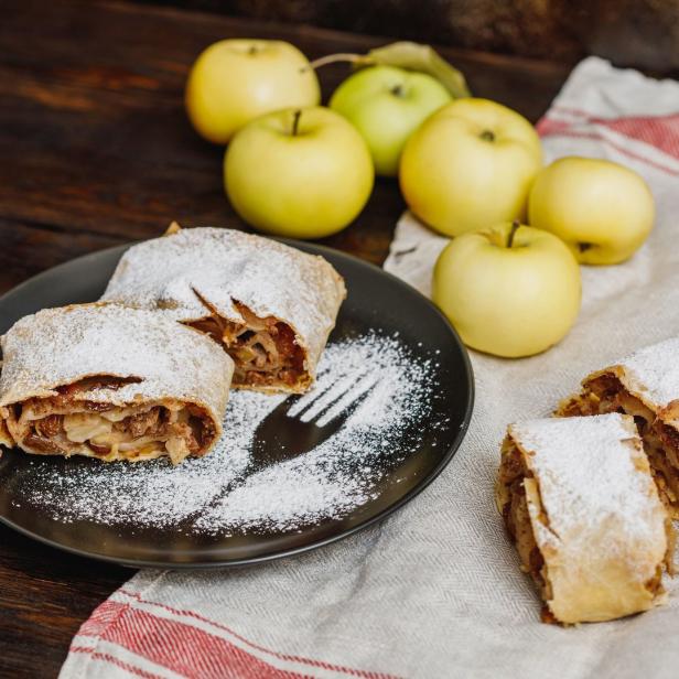 Apfelstrudel mit Puderzucker auf einem Teller mit Äpfeln im Hintergrund.