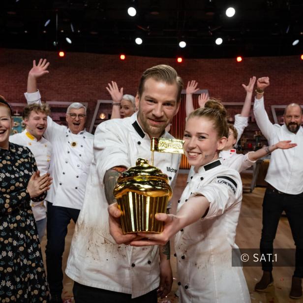 Zwei Bäcker präsentieren stolz eine goldene Cupcake-Trophäe, umgeben von jubelnden Kollegen.