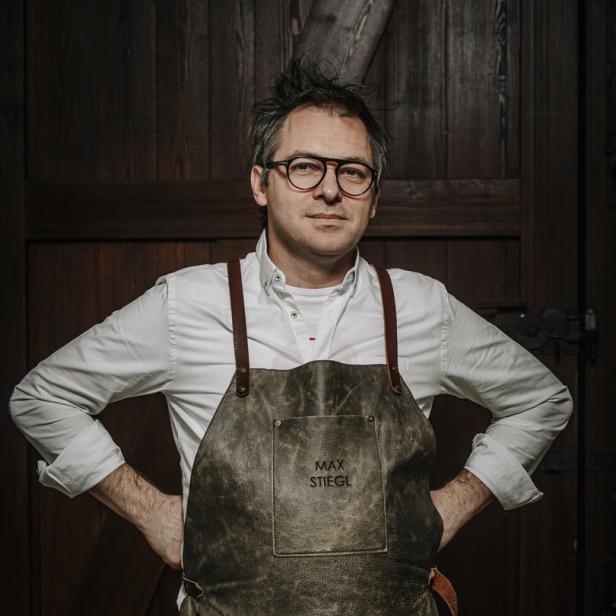 Max Stiegl mit Brille und Lederschürze vor einer Holztür.