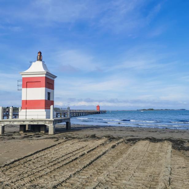 Ein rot-weiß gestreifter Leuchtturm steht an einem Strand mit einem langen Pier im Hintergrund.