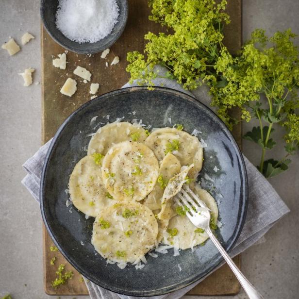 Ein Teller mit Ravioli, garniert mit Käse und frischen Kräutern, steht auf einem Holztisch.