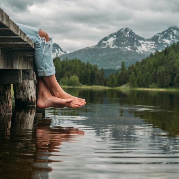 Eine Person sitzt barfuß auf einem Holzsteg an einem See mit Bergblick.