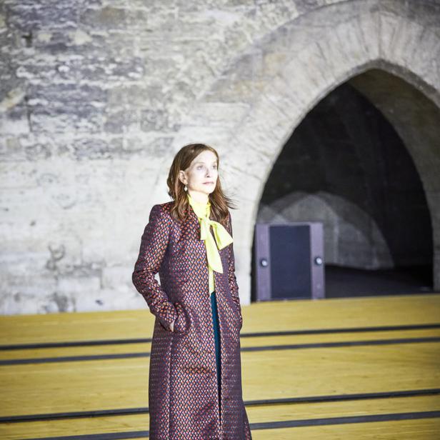 Isabelle Huppert posiert in einem gemusterten Mantel und türkisfarbenen Hosen vor einem Steingewölbe.
