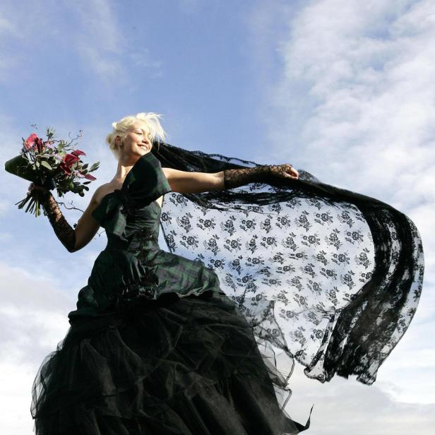Eine Frau in einem dunklen Kleid mit Schleier und Blumenstrauß steht auf Felsen vor einem Himmel mit Wolken.