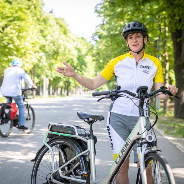 Eine Frau mit Helm steht neben einem E-Bike der Marke Cube auf einer Allee.