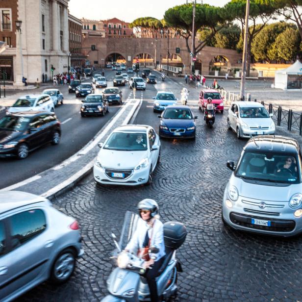 Eine belebte Straße in Rom mit Autos und einem Motorroller auf Kopfsteinpflaster.