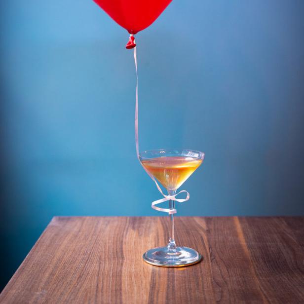 Ein roter Herzluftballon schwebt über einem Cocktailglas auf einem Holztisch.