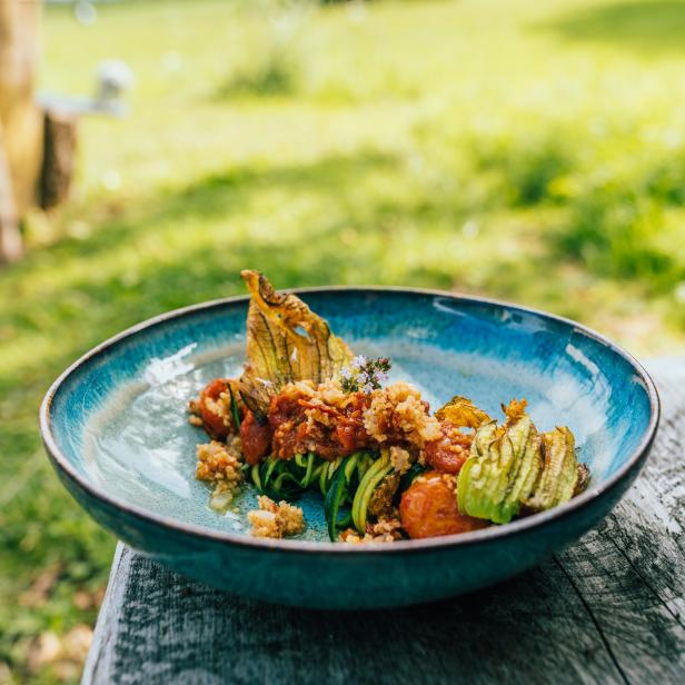 Ein Teller mit Zucchinigemüse, Tomaten und Zucchiniblüten steht im Freien auf einem Holztisch.