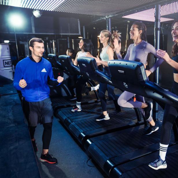 Eine Gruppe von Frauen trainiert lachend auf Laufbändern in einem Fitnessstudio, angeleitet von einem Mann.