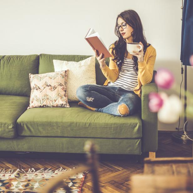 Eine Frau sitzt mit einer Tasse und einem Buch auf einem grünen Sofa.