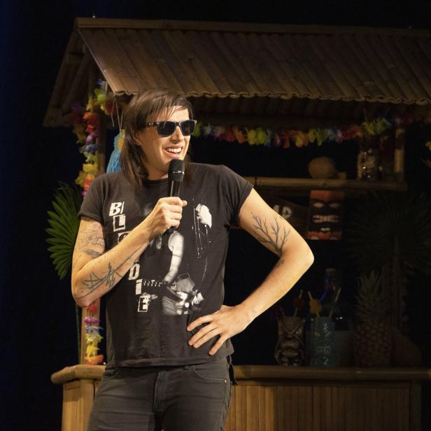 Eine Person mit Sonnenbrille und Tätowierungen tritt vor einer Tiki-Bar mit einem Mikrofon auf.