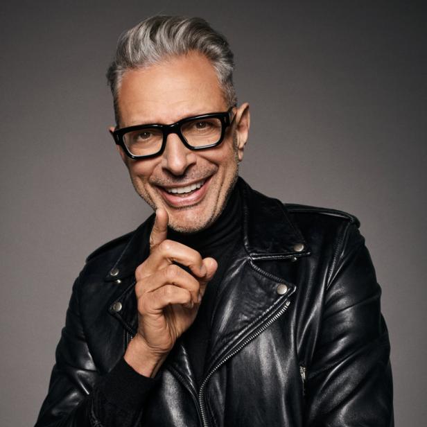 Jeff Goldblum lächelt in schwarzer Lederjacke und Brille, den Finger an die Wange gelegt.