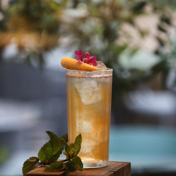 Ein Cocktail mit Eiswürfeln, einer Maiskolben-Garnitur und einer Blüte steht auf einem Holzsockel.