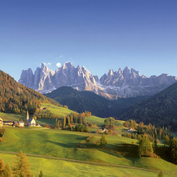 Blick auf ein Dorf in den Dolomiten mit einer Kirche und bunten Herbstwäldern.