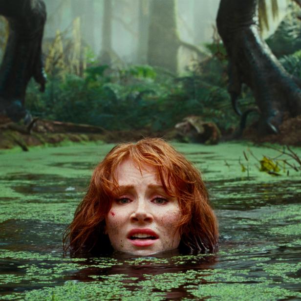 Eine Frau watet durch einen grünen Sumpf, während im Hintergrund Dinosaurierfüße zu sehen sind.